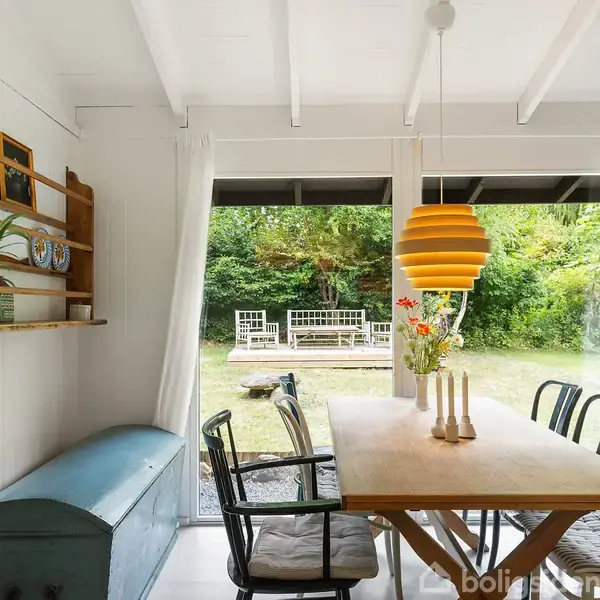 Et spisebord med stole omkring står i et lyst rum med hvide vægge og synlige loftsbjælker. En gul lampe hænger over bordet, og udenfor ses en grøn have.