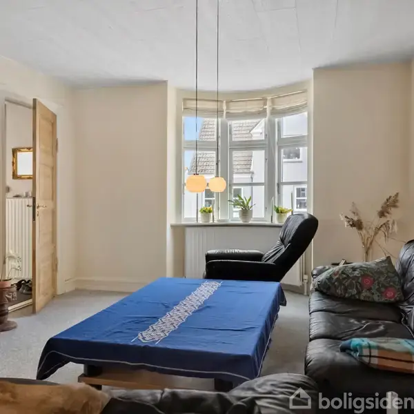 Stue med sort lædersofa omkring et sofabord dækket af en blå dug. En lænestol vender mod et stort vindue med planter. Der hænger lamper i vinduet.