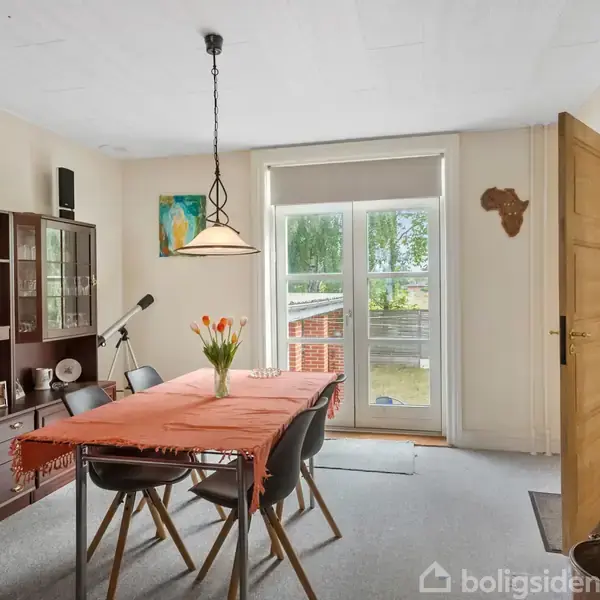 Et spisebord med en orange dug står i en lys spisestue, omgivet af seks stole. En vase med blomster er på bordet, og en stor reol med dekorationer står langs væggen. Dobbeltdøre fører ud til en have.