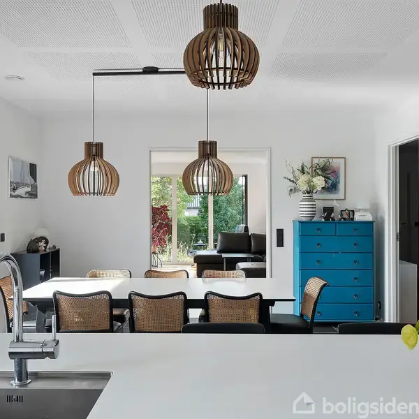 Køkkenbord med moderne vandhane står i forgrunden. Over bordet hænger tre dekorative lamper. I baggrunden ses et spisebord, blåt skab og udsigt til en terrasse samt have.