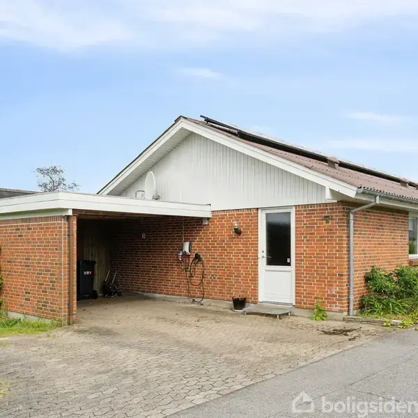En lav, rød murstensvilla med en carport og sort tag med solpaneler står på en brolagt indkørsel, omgivet af hække og buske under en klar, blå himmel.