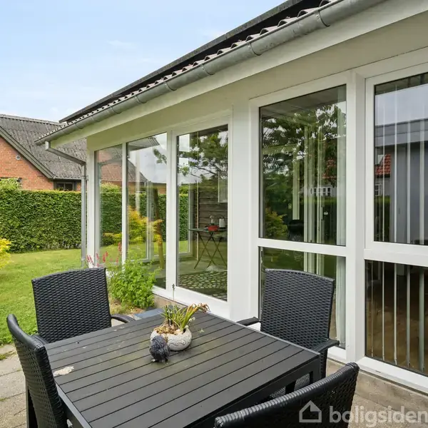 Et sort udendørs bord står på en flisebelagt terrasse ved siden af en hvid udestue. Der er fire sorte stole omkring bordet. En grøn græsplæne omgiver området.