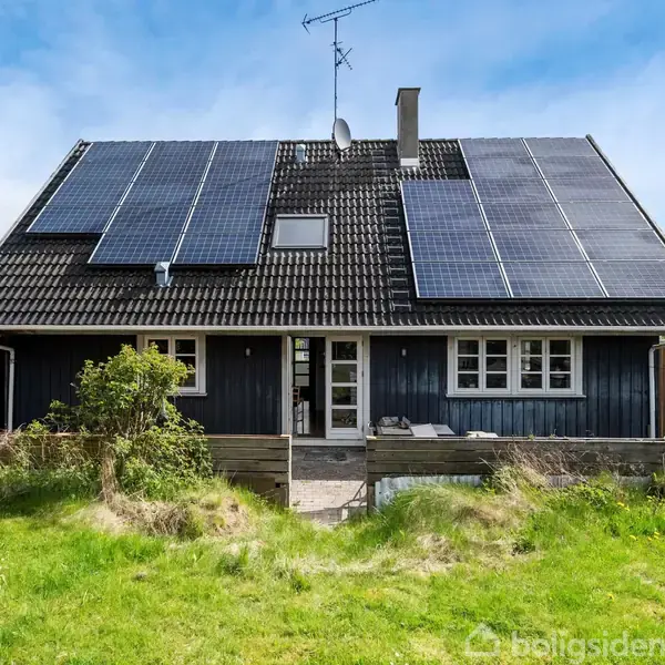 Hus med solpaneler monteret på taget, omgivet af grøn græsplæne og hæk. Blå himmel i baggrunden.