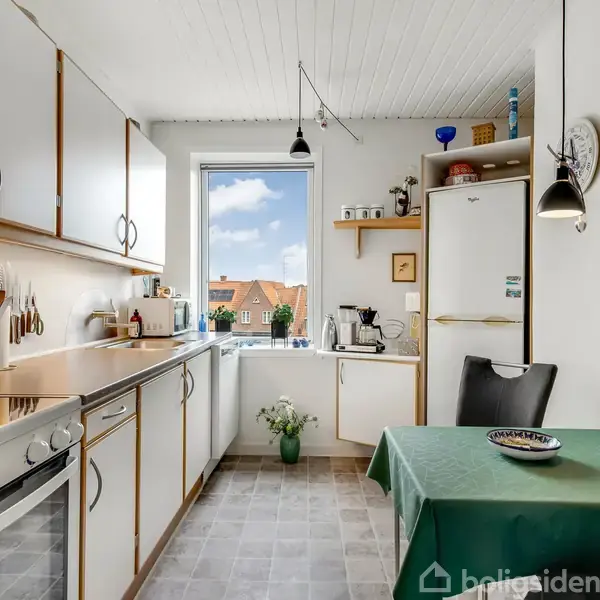 Køkken med hvide skabe og en ovn til venstre, står stille. Et lille bord med grøn dug og to stole er til højre. Vindue centralt med udsigt til hustage.