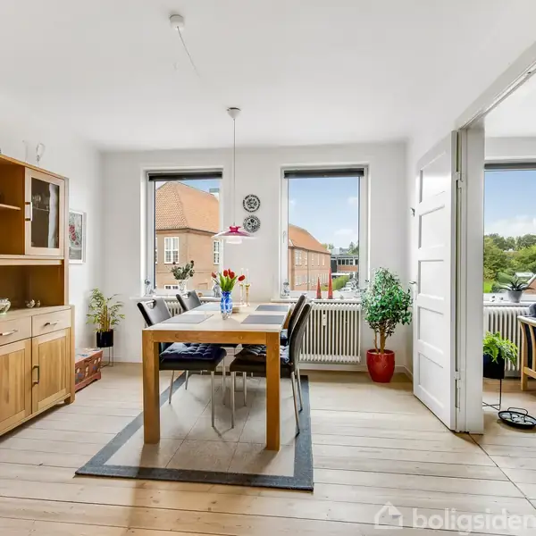 Spisebord med stole står i en lys spisestue. Et vitrineskab står til venstre, mens en sofa er synlig i tilstødende rum. Vinduernes udsigt viser bygninger og træer.