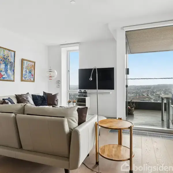 En lys stue med en beige sofa foran et fladskærms-tv, placeret ved siden af en udendørs balkon med udsigt. Rummet har lamper, farverige billeder på væggene, og et rundt sofabord.