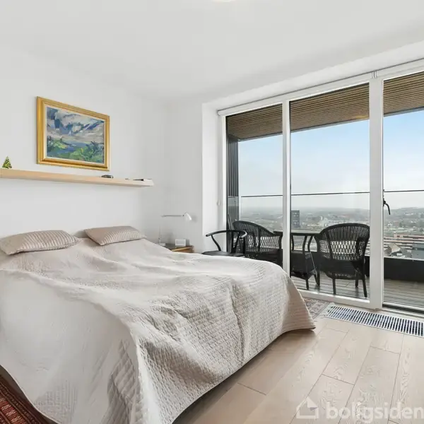 En seng står i et lyst soveværelse, dækket med hvidt sengetøj. Et maleri hænger på væggen, og en stor glasdør fører til en balkon med udsigt og siddemøbler.