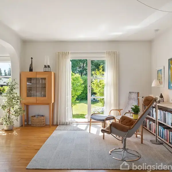 En lys stue indeholder en lænestol, bogreol og tæppe, med udsigt til en grøn have gennem store vinduer. Rummet er indrettet med minimalistisk stil og naturligt lys.