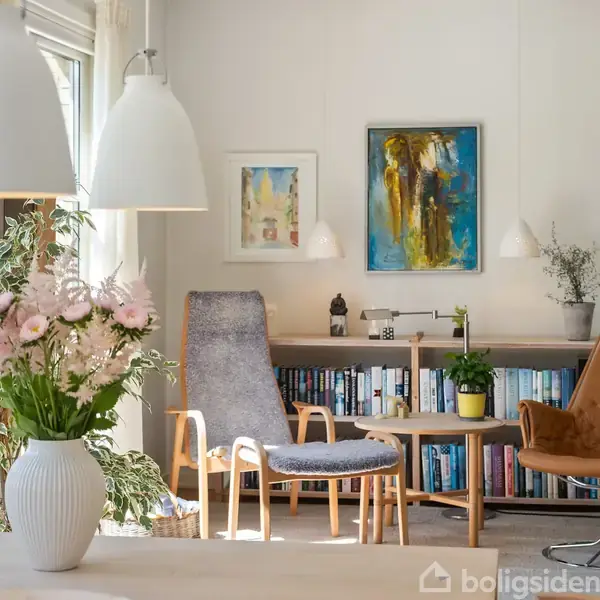 En stue med to stole står ved siden af et lille bord foran en reol fyldt med bøger. Vaser med blomster og kunstværker pynter rummet.