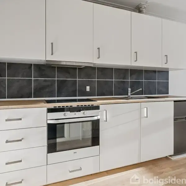 Et moderne køkken med hvide skabe, en indbygget ovn og en keramisk kogeplade. Skuffesektioner er til venstre, og en vask er placeret på højre side. Baggrunden har sorte fliser.