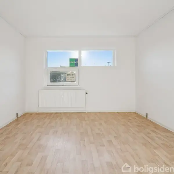 Et tomt rum med lyst trægulv, hvide vægge og et vindue med radiator under. Vinduesudsigten viser blå himmel og tagene af bygninger udenfor.