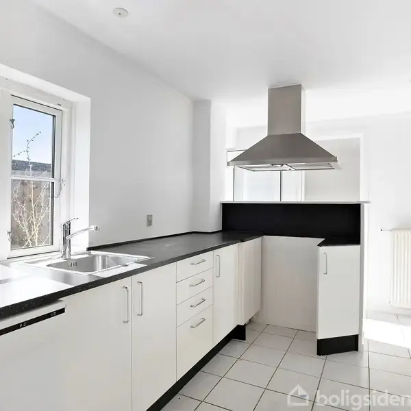 Et moderne køkken med hvide skabe og sort bordplade. En stor emhætte er centreret over kogepladsen. To vinduer giver naturligt lys. Radiator ved væggen.