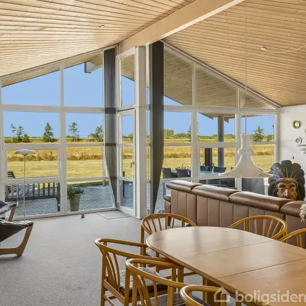 Stue med store vinduer og skråt loft, der lukker naturligt lys ind. Et spisebord står centralt omgivet af stole; i baggrunden ses en sofa og fladskærms-TV. Udsigt til landskab.