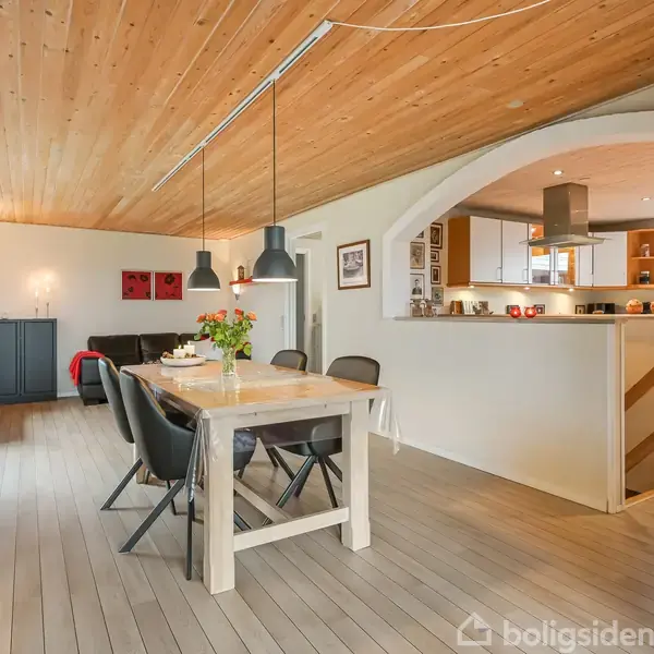 Spisebord med stole står i midten af en lys spisestue. Træloft og gulv skaber en varm atmosfære. Køkken ses gennem en bueåbning i baggrunden. Vægdekorationer og lamper tilføjer hygge.