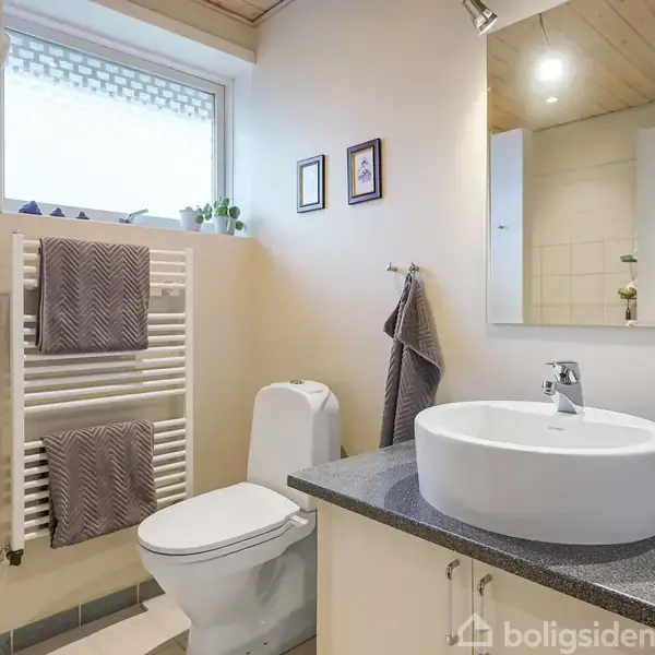 Håndvask med rund skålform står på en grå bordplade, omgivet af hvidt skab. Ved siden af ses et toilet og håndklæder på vægmonteret holder. Vindue og spejl bagved.