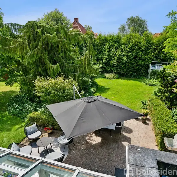 Stor parasol står over havemøbler på en terrasse. Omkring er en velplejet have med grønne buske, høje træer og en græsplæne. En grill er placeret ved siden af møblerne.