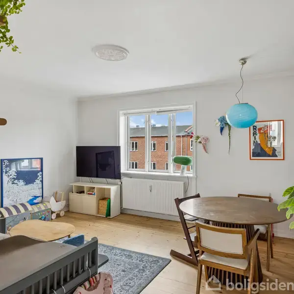 Et lyst stueområde med et rundt spisebord og stole. Et tv står på en tv-bænk ved et vindue. Planter og bøger pryder hylder i et roligt hjemmemiljø.