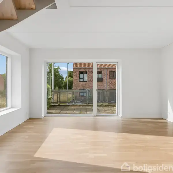 Lys stue med trægulv, store vinduer og glasdøre, der fører ud til en terrasse. I baggrunden ses en have og røde murstenshuse. Trappe i venstre hjørne.