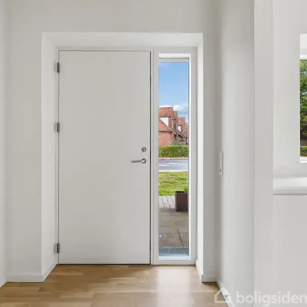 Dør med sidevindue står i en lys entre med trægulv. Udenfor ses en græsplæne og røde bygninger. Til højre er et køkkenvindue med udsigt til grønne træer.