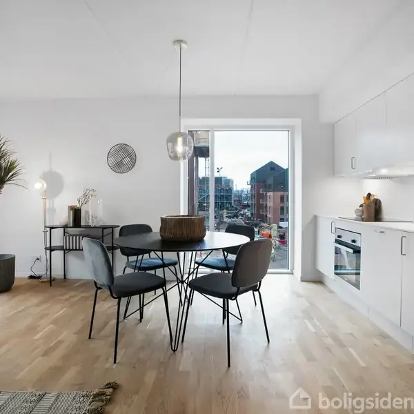 Rundt bord med fire stole står i en lys stue-køkken kombination. Køkkenet har hvide skabe, og store vinduer giver udsigt til bymiljø. En plante står i hjørnet.