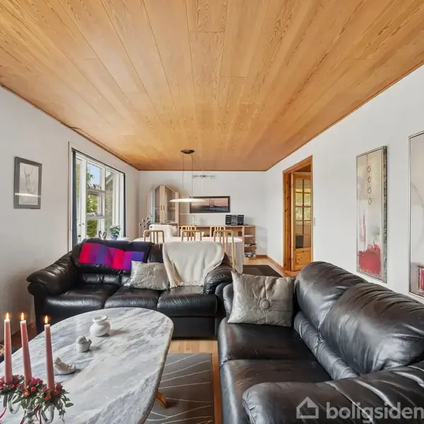 Stue med sorte lædersofaer omkring et sofabord med lysestager. Rummet har trægulv og loft, vinduer til venstre, malerier på væggen, og åbner ind til et spisekøkken.