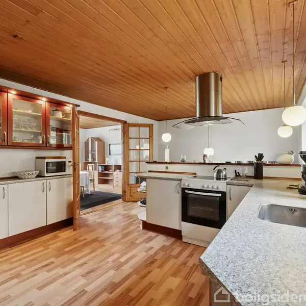 Køkken med træloft og trægulv. Køkkenskabe i hvid og træfarve. En moderne ovn og komfur er centralt placeret. Bordplader af granit. Lyse lamper hænger fra loftet.