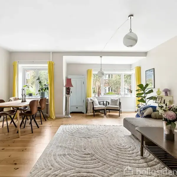 En stue med et spisebord til venstre, sofa og sofabord til højre. Store vinduer med gule gardiner slipper lys ind. Vægkunst og grønne planter pryder rummet.