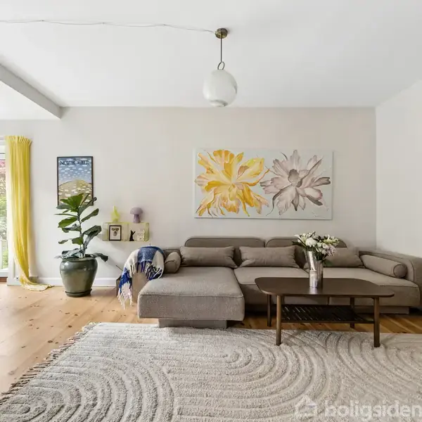Sofa står i lys stue ved hvide vægge og trægulv, pyntet med blomsterbilleder. To vinduer omgivet af gule gardiner, planter, og bogreol skaber en hyggelig atmosfære.