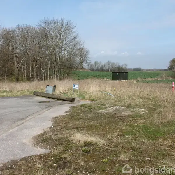 En ubrugt landevej ender ved et græsområde med træer i baggrunden. En lille betonblok med skilt står ved vejsiden. Grønt marker strækker sig mod horisonten.