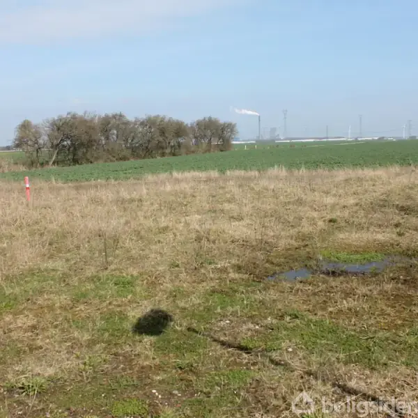 En åben mark med græs og ujævnt terræn ligger stille. I baggrunden er der små træer og marker, mens industrielle strukturer kan ses i horisonten under en blå himmel.