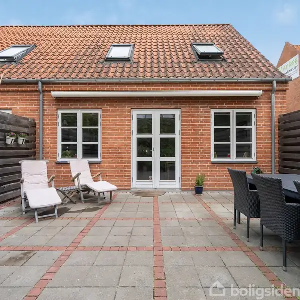 Rød murstensbygning med terrasse, hvor der står to solsenge til venstre og et bord med stole til højre, omgivet af hegn. Terrassefliser danner gulvet i en solrig gårdhave.