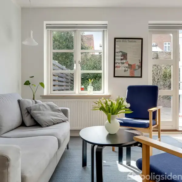 Grå sofa med puder står i en stue ved et rundt bord med blomster. Et blåt tæppe ligger på gulvet, og vinduer viser en have udenfor. En plakat på væggen.