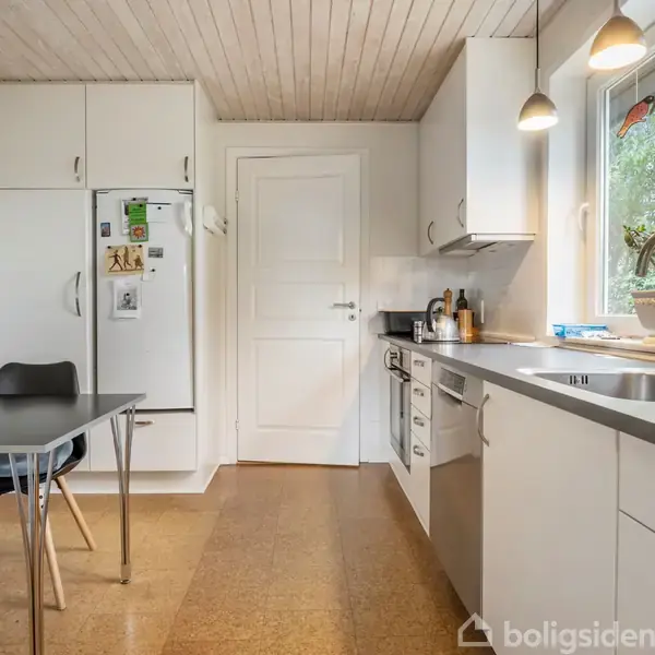 Køkken med hvidt interiør, spisebord til venstre, køleskab mod væggen, og en vask ved vinduet til højre. Vinduet viser grøn udsigt udenfor.