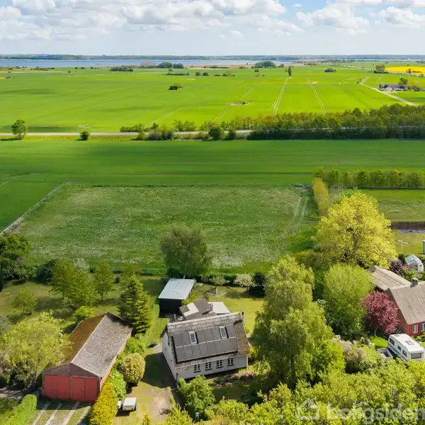 Et landhus med gråt tag ligger i en frodig have omgivet af træer. To tilstødende bygninger ses, og marker strækker sig ud til horisonten under en blå himmel.