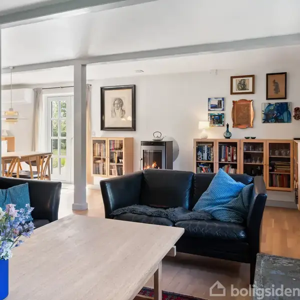 Sort sofagruppe med blå puder står foran et træbord med blomster i en vase. Rummet er lyst, med hylder og kunst på væggen samt en spiseplads i baggrunden.