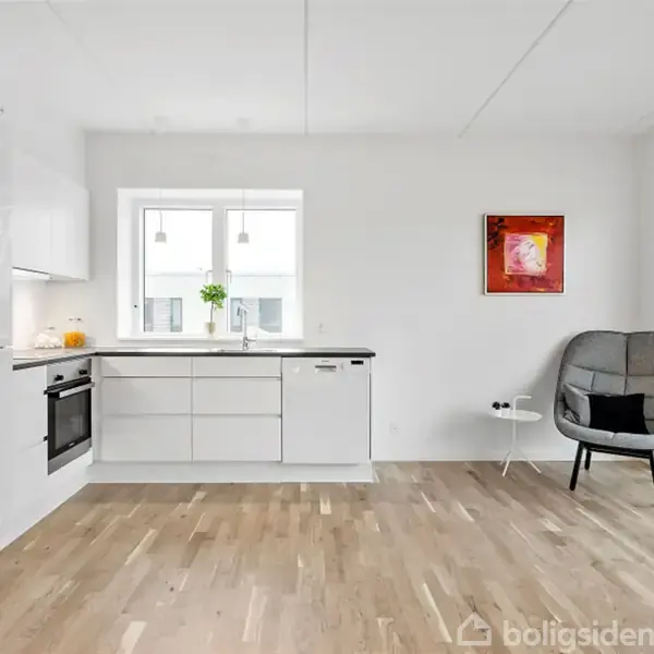 Et moderne køkken med hvide skabe og indbygget ovn. En stor stol står ved en væg med et farverigt billede. Trægulv og store vinduer giver lys og rummelighed.
