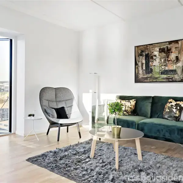 Stue med grøn sofa står på gråt tæppe ved siden af rundt sofabord. Til venstre en lænestol med pude. Væggen har kunstmaleri; glasdør fører til altan.