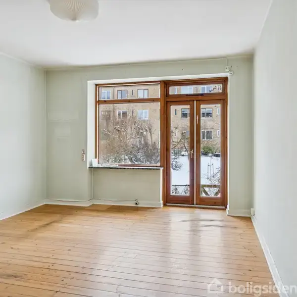 Et tomt rum med trægulv har en stor vinduesdør, der fører ud til en sneklædt have. Væggene er lyse, og en radiator står under vinduet.