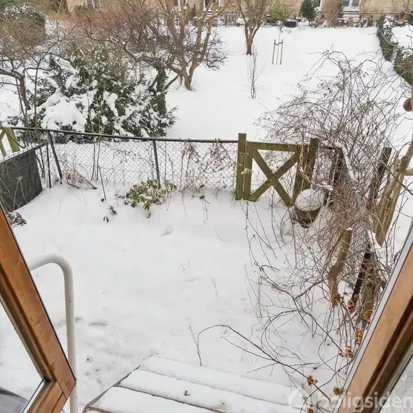 En snedækket have set fra en døråbning. Sne dækker græsset og træerne. En hegnsporte står åbent. Alt ser vinterpræget og stille ud.