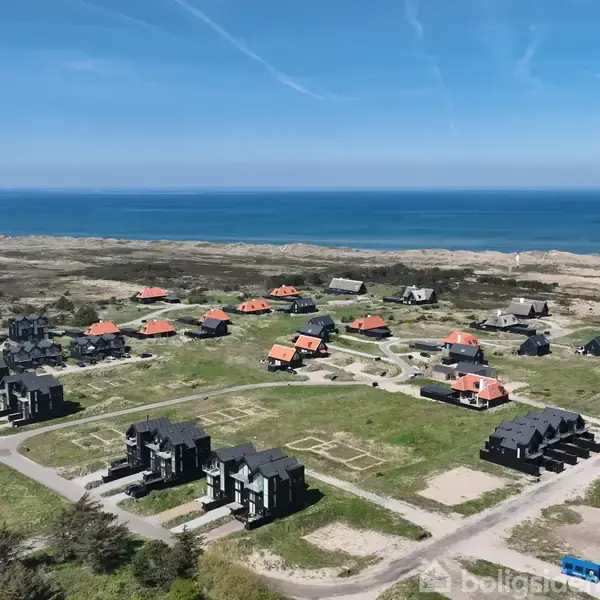 Flere moderne huse med mørke tage står i et boligområde nær kysten. De er omringet af græsarealer og stier, med havet i horisonten under en klar blå himmel.