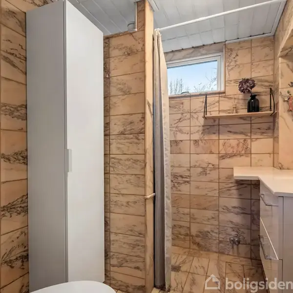 Et badeværelse med beige marmorerede fliser; en hvid skabslåge står til venstre. En bruseradskillelse er synlig, med et lille vindue ovenfor. En hylde med vaser og pynt genstande hænger på væggen.