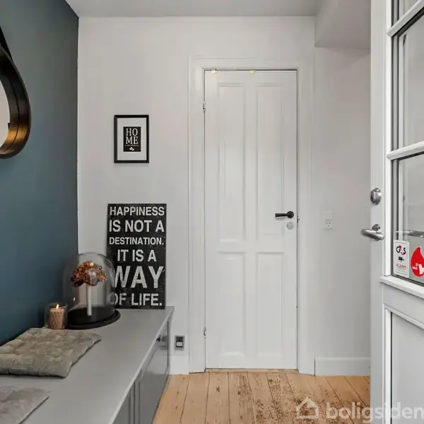 En entre med en bænk og puder langs en væg. Der hænger et rundt spejl over bænken. Tekstplakat siger: "HAPPINESS IS NOT A DESTINATION, IT IS A WAY OF LIFE." En dør og trægulv.