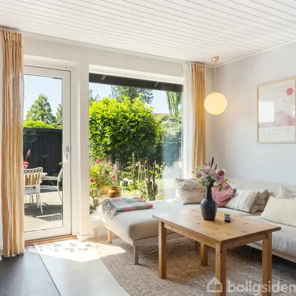 Stue med en lys sofa og sofabord dekoreret med blomster, set mod en dør førende til en solrig have. Væggen har kunstplakater, og pejsen er tændt.