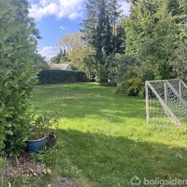 Fodboldmål stående på en grøn græsplæne omgivet af træer og buske under en blå himmel med skyer. En blå krukke med planter står i forgrunden.