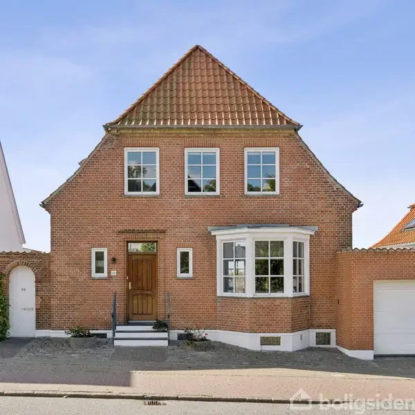 Et rødt murstenshus står stille på en gade, med et trædør og flere hvide vinduer. Husets højre side grænser til en garage. Fladen er omgivet af en asfalteret vej.