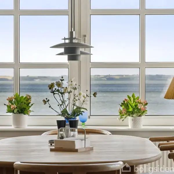 Et rundt træbord står foran store vinduer med udsigt til havet. På bordet er en vase med blomster, og i vinduet er der planter og en lampe.