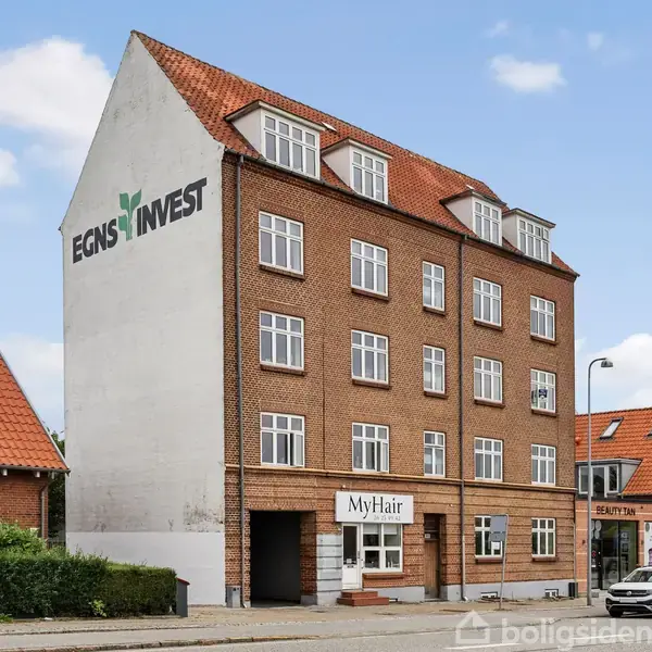 En brun murstensbygning med skiltet "EGNSINVEST" på siden står i en bymæssig gade. I stueetagen er der en frisørsalon med navnet "MyHair". To biler er parkeret foran.