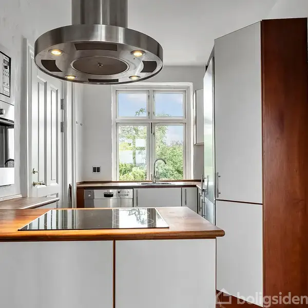 Et moderne køkken med en kogeø og emhætte over, indbyggede ovne i væggen. Store vinduer giver udsyn til en grøn have, og hvide skabe dominerer rummets design.