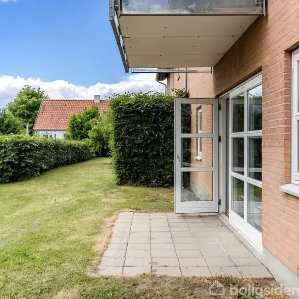 Dør åbner mod have med flisebelægning i forgrunden og en bil parkeret på græsplæne. Hæk og hus i baggrunden under en blå himmel med skyer.