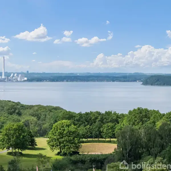 En sø danner midtpunktet med en industriel struktur i baggrunden. Grønt landskab med træer og en rødbygget bolig fylder forgrunden under en skyfri blå himmel.